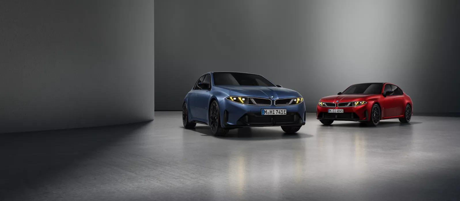 Két BMW 3 M modell, megvilágított veserácsokkal, világosbarna felületen parkolva.